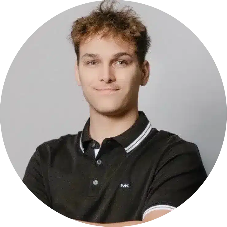 A young man with short, light brown hair is standing against a plain background, arms crossed and wearing a black polo shirt with white trim—a confident member of Our Team looking directly at the camera.