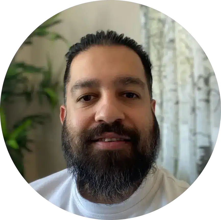 A bearded man with dark hair, part of Our Team, is smiling at the camera in a white shirt. Green plants and a curtain with a tree pattern create a welcoming backdrop.