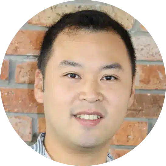 A man with short black hair smiles slightly in front of a brick wall background, reflecting the warm and approachable vibe of a Chiropractor Coquitlam professional.